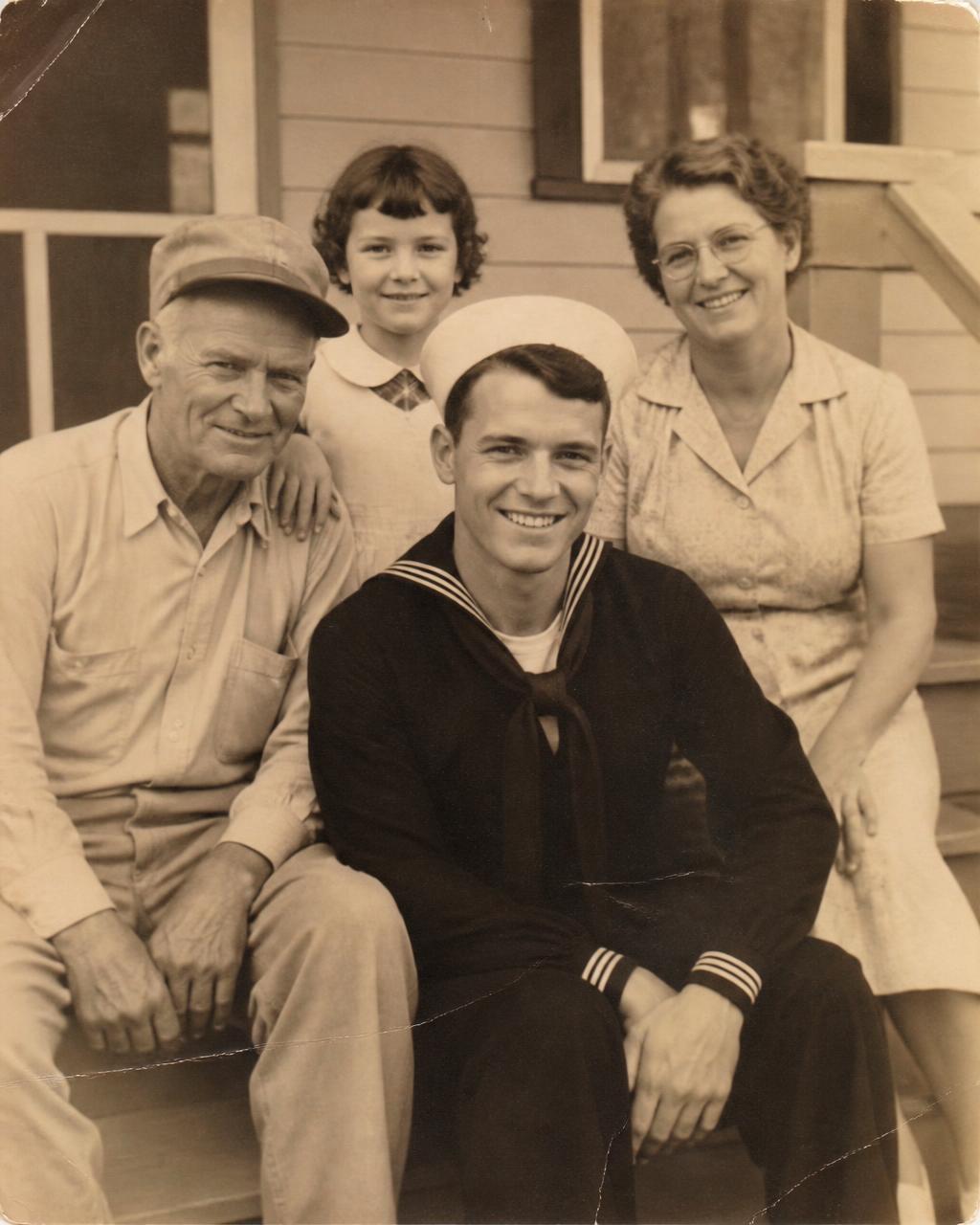 Sepia family photograph of a young sailor seated with his parents and younger sister on a porch.